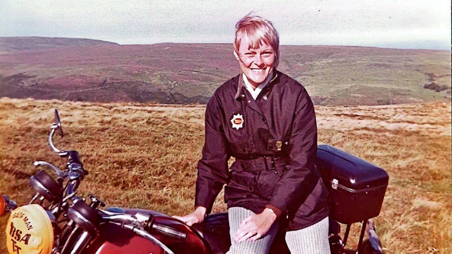 Cherry Allen, sat on a motorbike smiling on the mountain in the Isle 0of Man, she has light hair and a firnge.