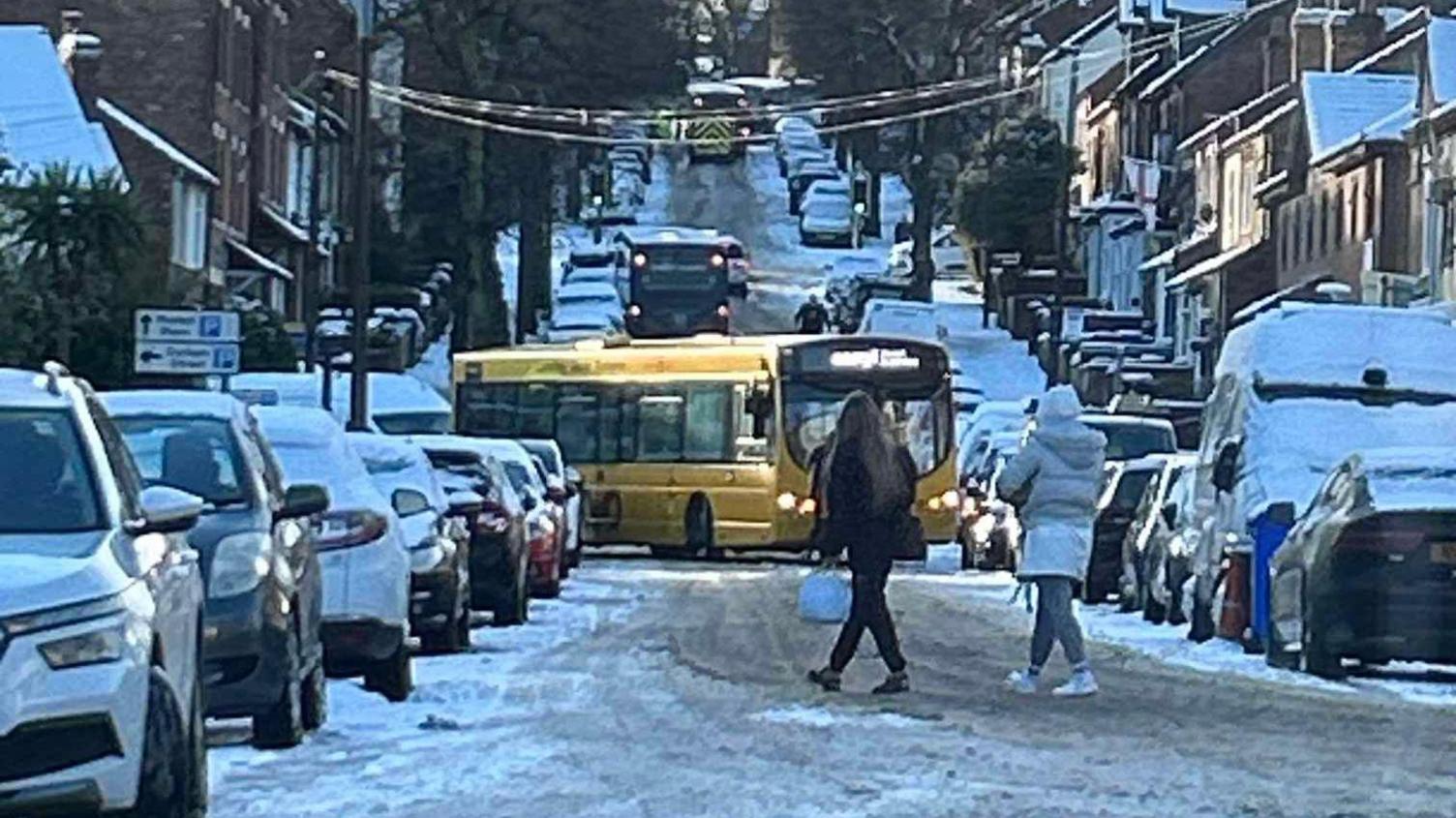 A bus at a sideways angle with snow on the ground. Vehicles are parked either side of the road