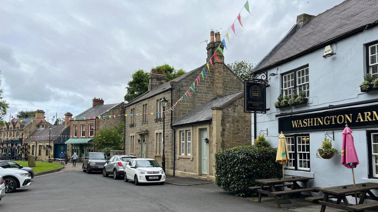 A row of stone buildings face into a village square. The nearest building is a pub, which painted blue and has a sign reading "Washington Arms" on the front. Colourful bunting is strung up from lamppost to lamppost.