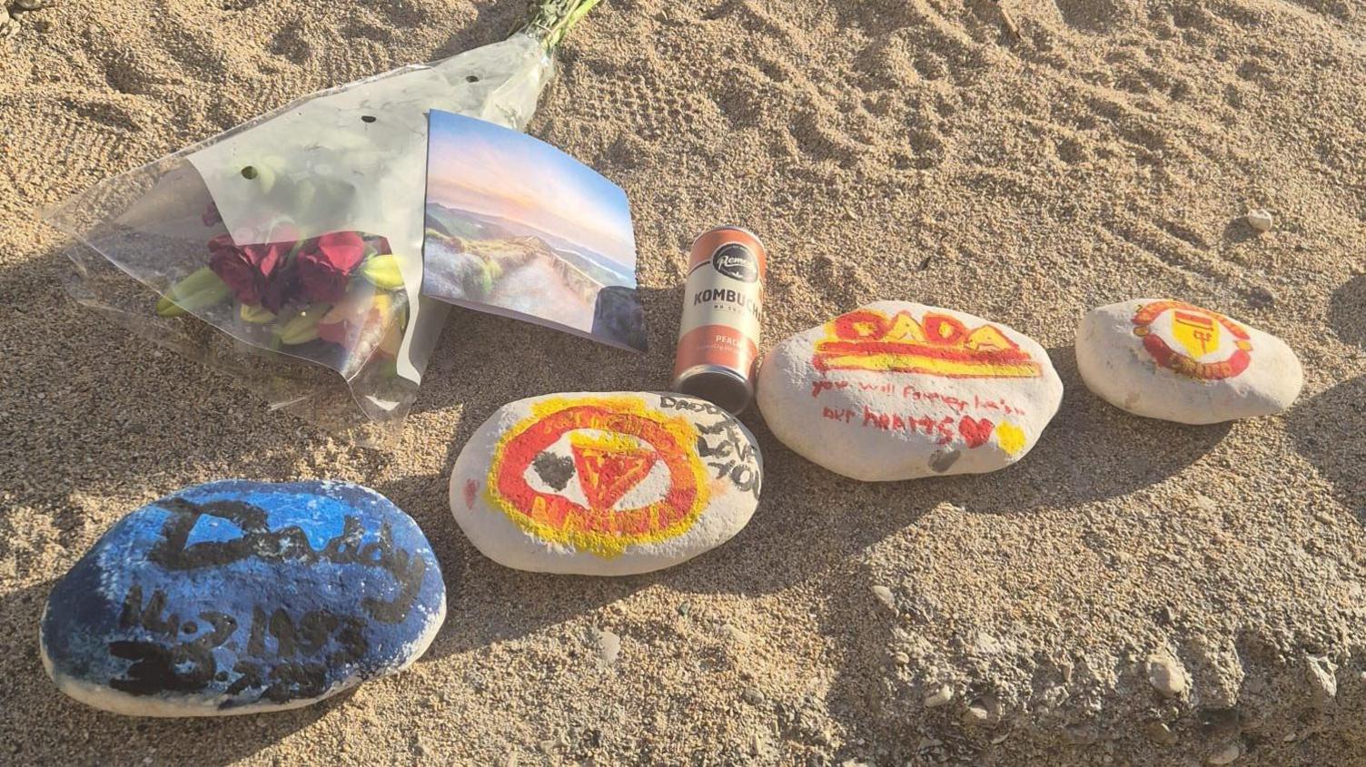 A row of four painted pebbles lie on the beach, alongside an orange drinks can, photograph and a bunch of flowers.