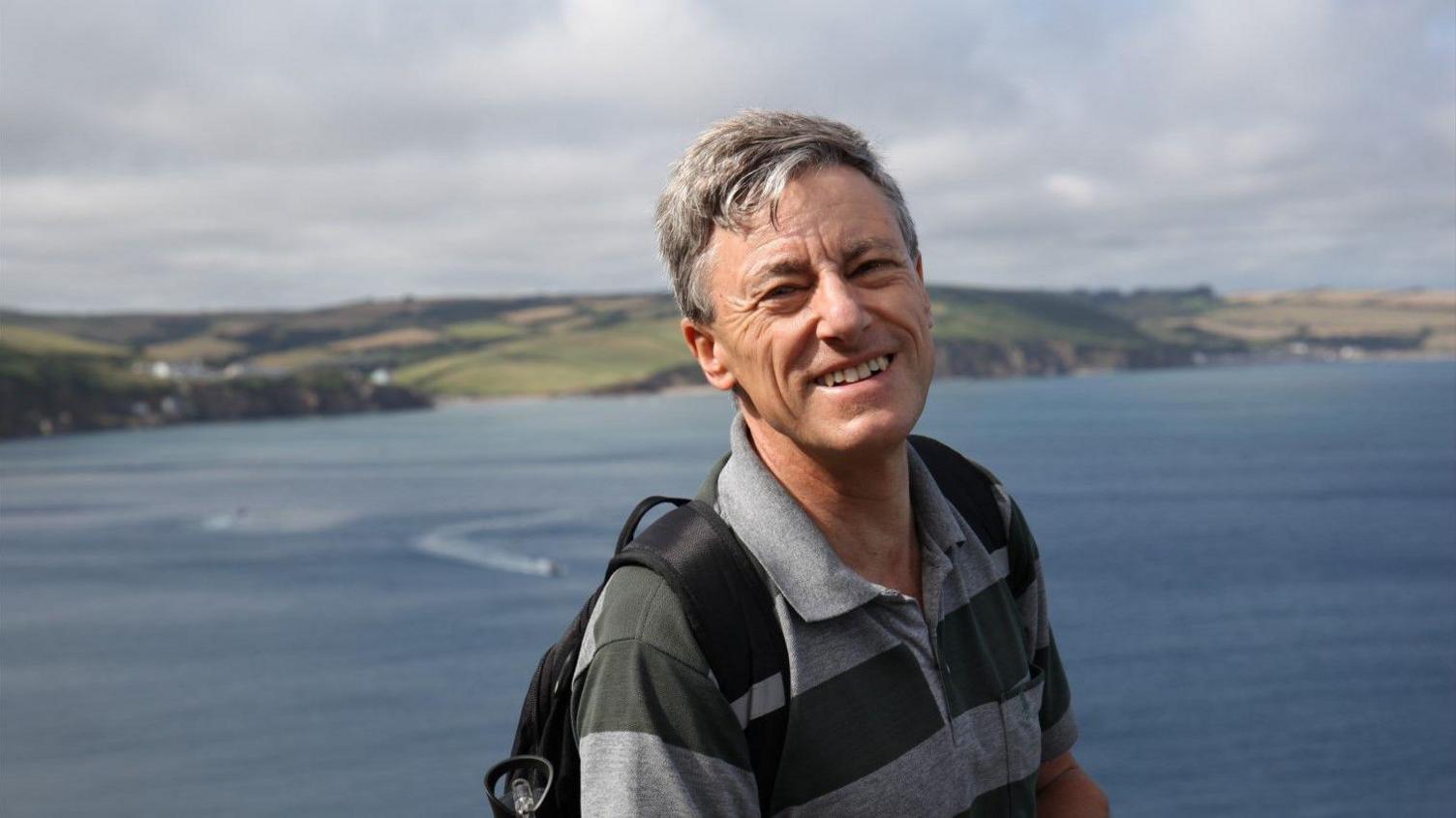 Robert Clark smiling at the camera wearing a grey and black striped polo and dark coloured backpack with short white hair.  A view of a body of water and coastline in the distance can be seen in the background.