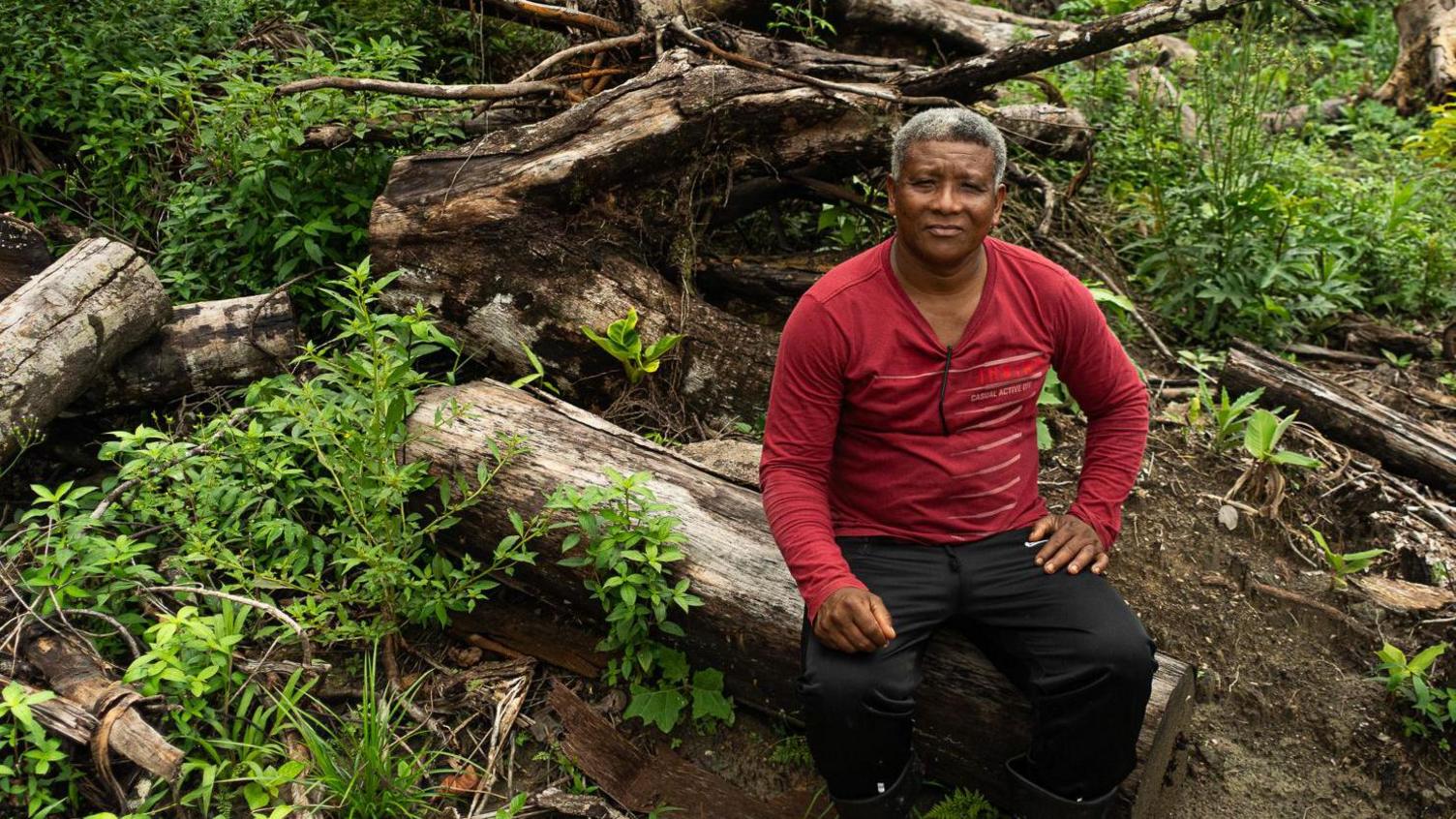 Claudio Verequete sits on a felled tree, wearing a red jumper. He has short grey hair and is looking at the camera