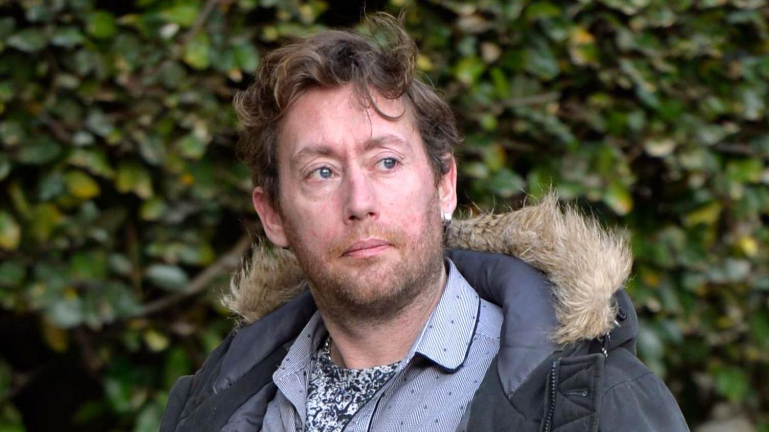 A man in his late 30s with wavy brown hair, a blue parka and a blue shirt outside the High Court in Glasgow. There are bushes in the background of the image and he looks to the right of where the camera is.