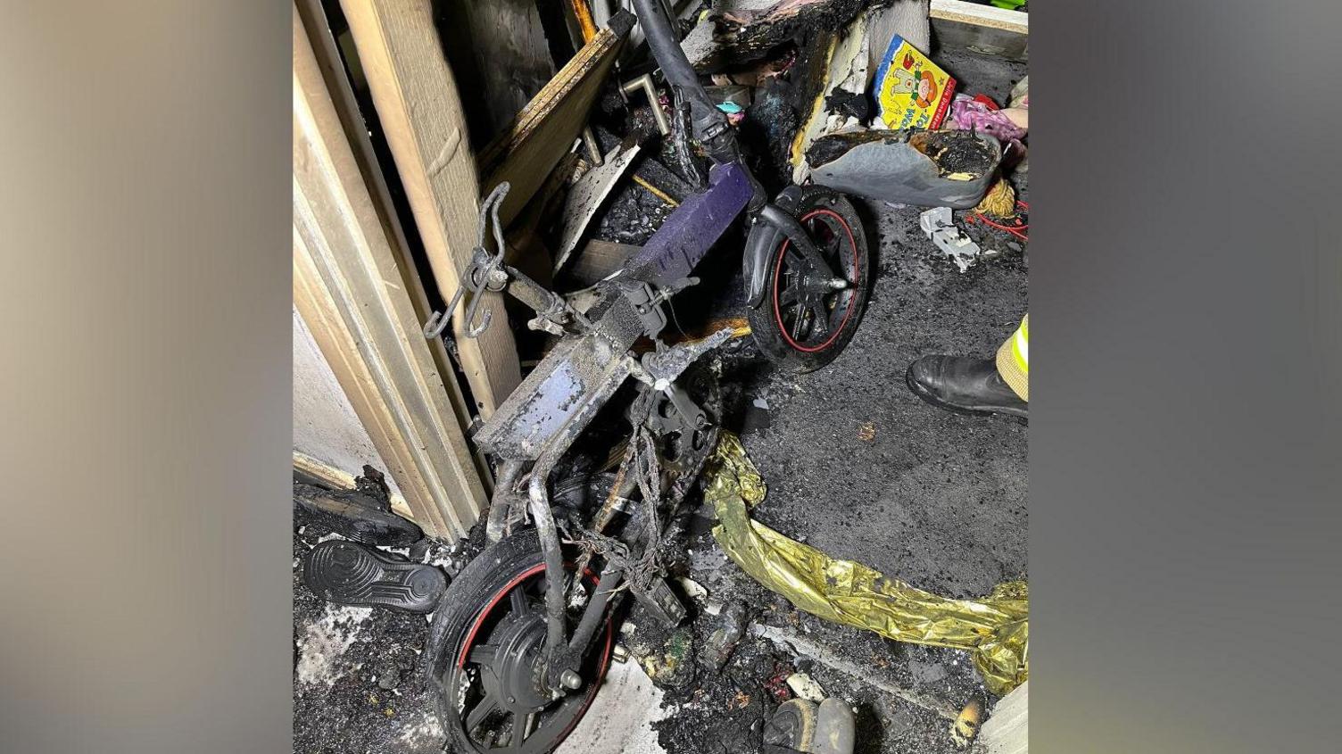 Fire service image of a charred small wheeled bicycle leaning against a smoke blackened wall in a domestic hallway