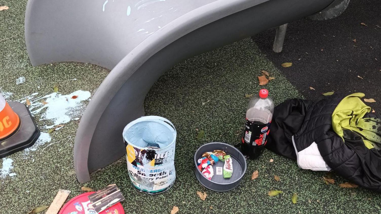 A cone at the bottom of the slide which has blue paint on it. There is a red celebrations lid with a pouch of tobacco on top to the right of the slide. There is also a open pot of blue paint next to it, a bowl with wrappers in, a bottle of fizzy drink and coat. 