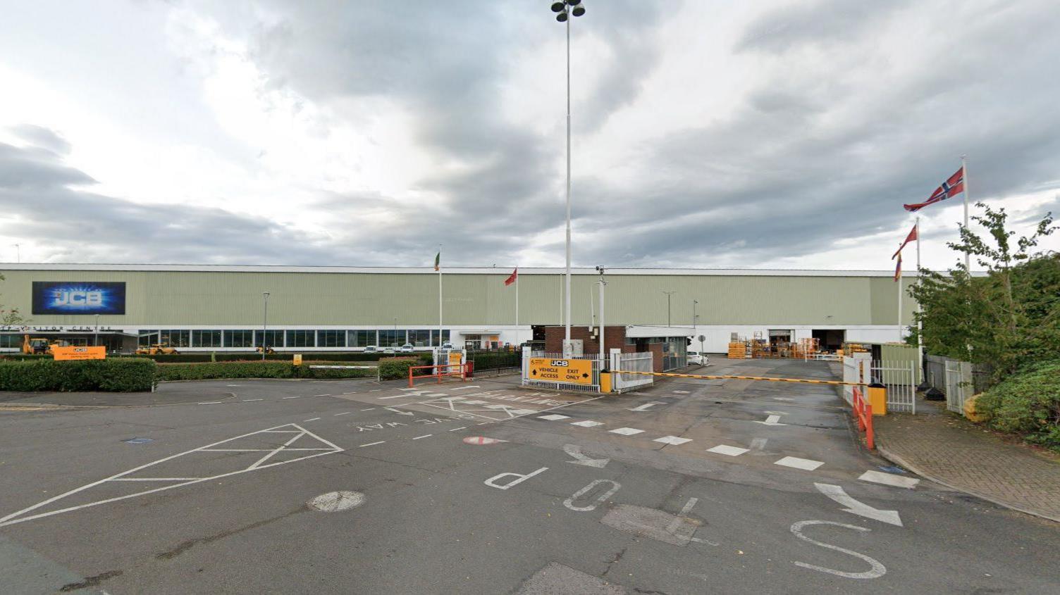 The entrance to the JCB world headquarters in Rocester, Staffordshire. A large, green warehouse-type building with a blue sign with JCB written on it in white letters. 