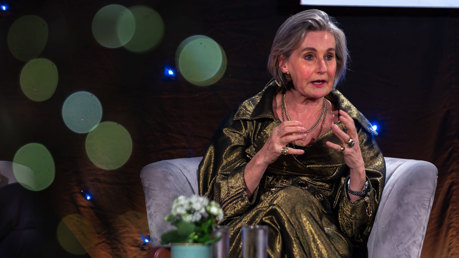 A woman with shoulder-length grey hair, gestures with her hands as she sits on a grey chair on a stage. She is wearing a sparkling gold outfit, with gold rings and necklaces.