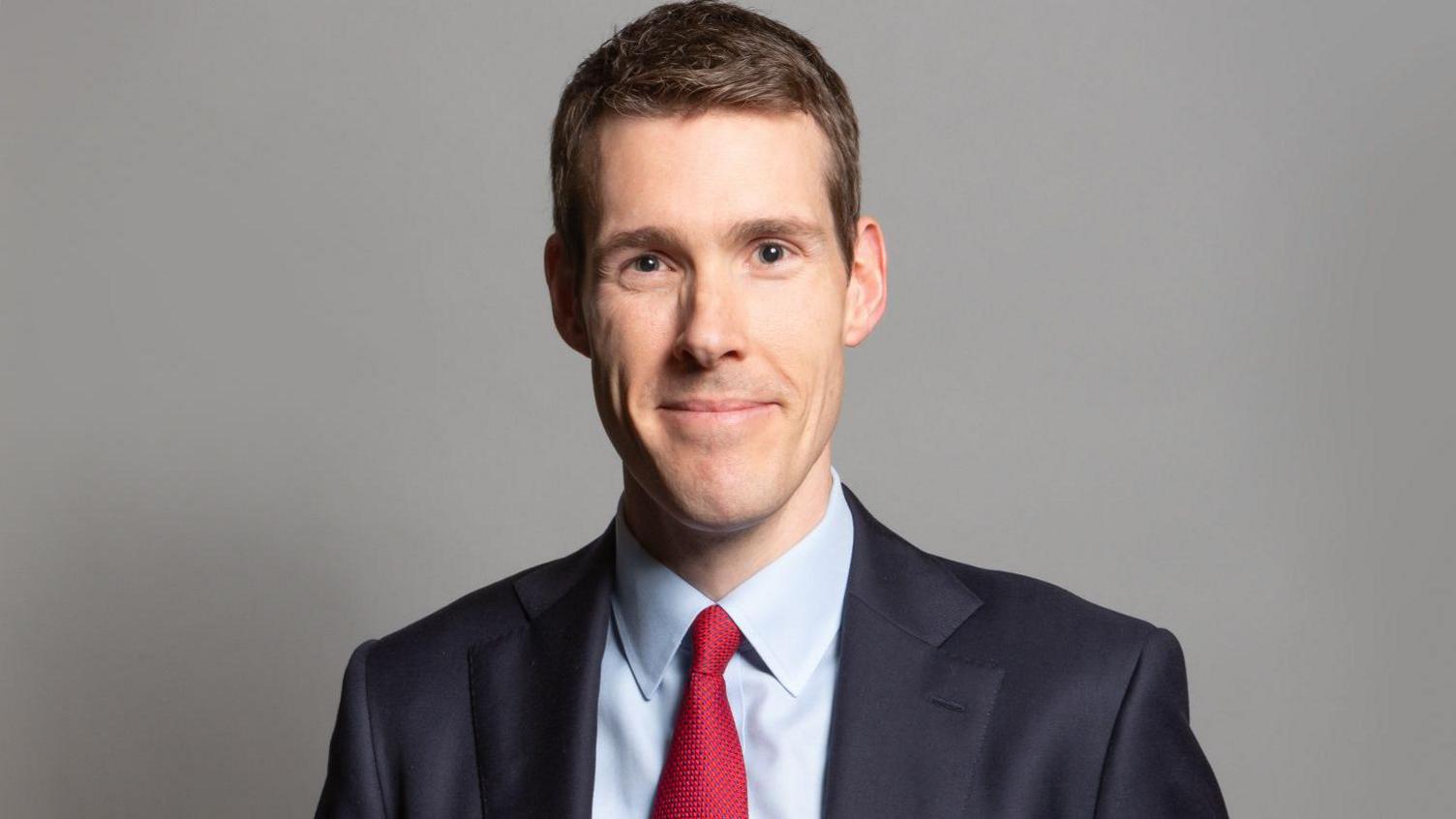 A professional head and shoulders photo of a man with short blonde hair and wearing a black suit with a red tie. He's standing against a grey background and smiling at the camera.
