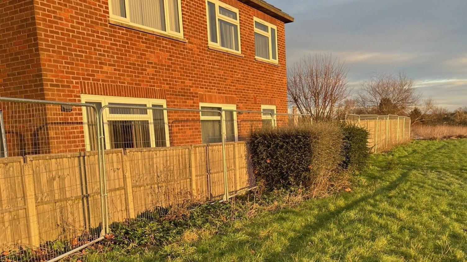 A two-storey home made from red bricks which is situated just behind a wooden picket fence. There is now metal heras fencing running parallel to the wooden fence, which is very tall and blocks the view from the windows. On the right side of the fencing there is a lush green field.