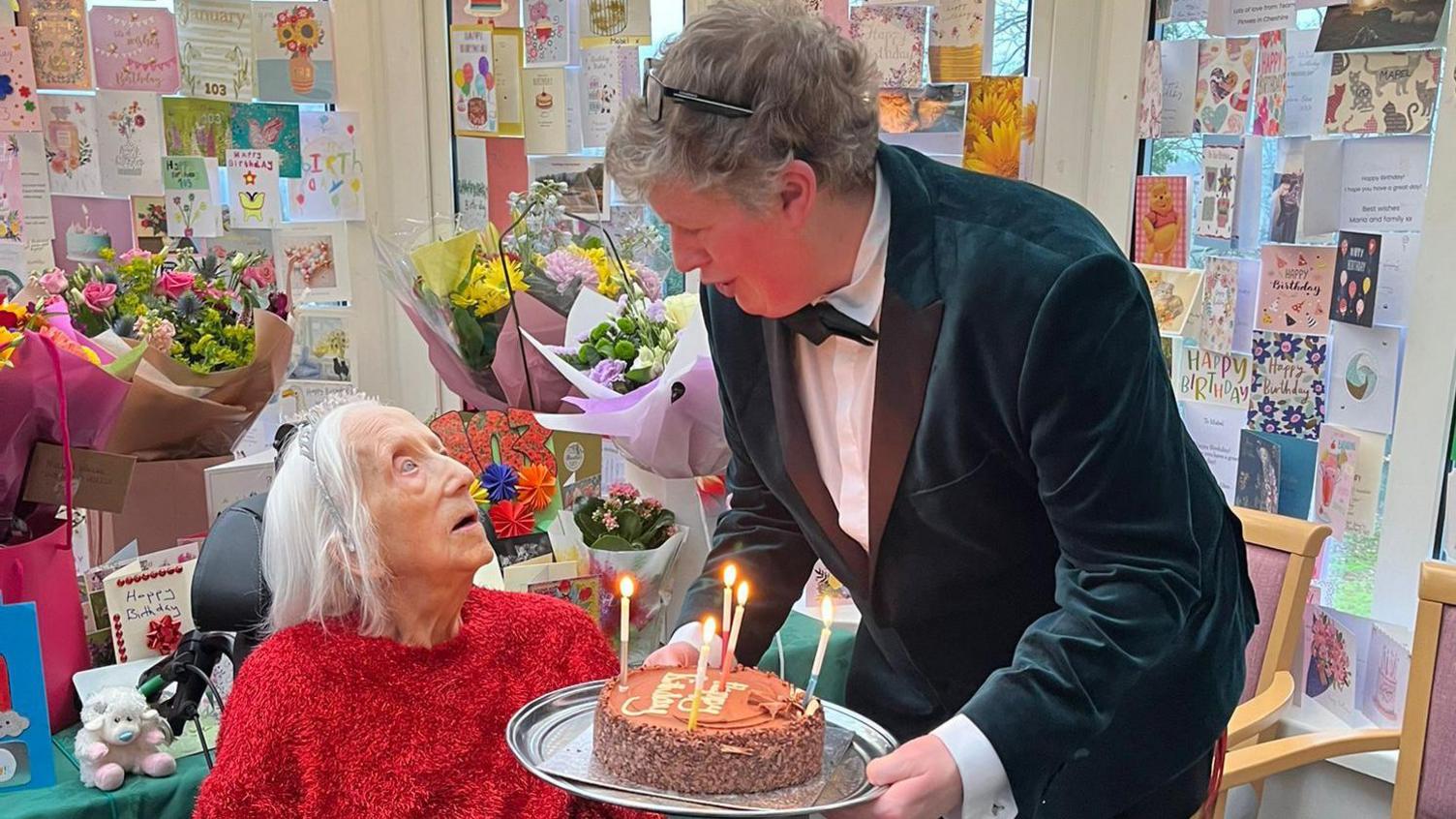 An image of a man in a suit presenting an elderly lady with a birthday cake