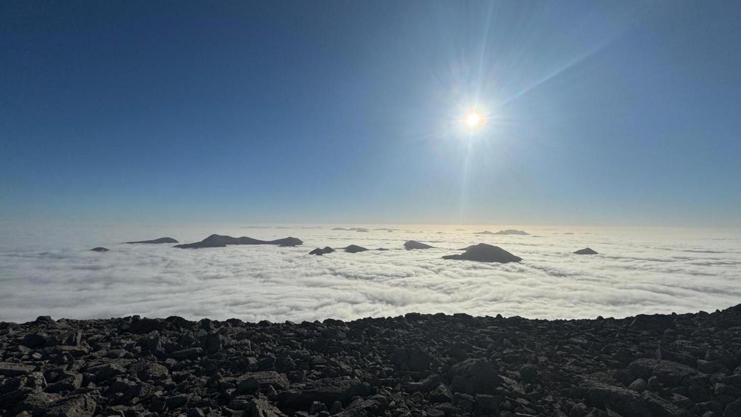 The ground is rocky in the foreground and beyond that is white cloud all the way to the horizon. Peeking up from the cloud are the tops of mountains.