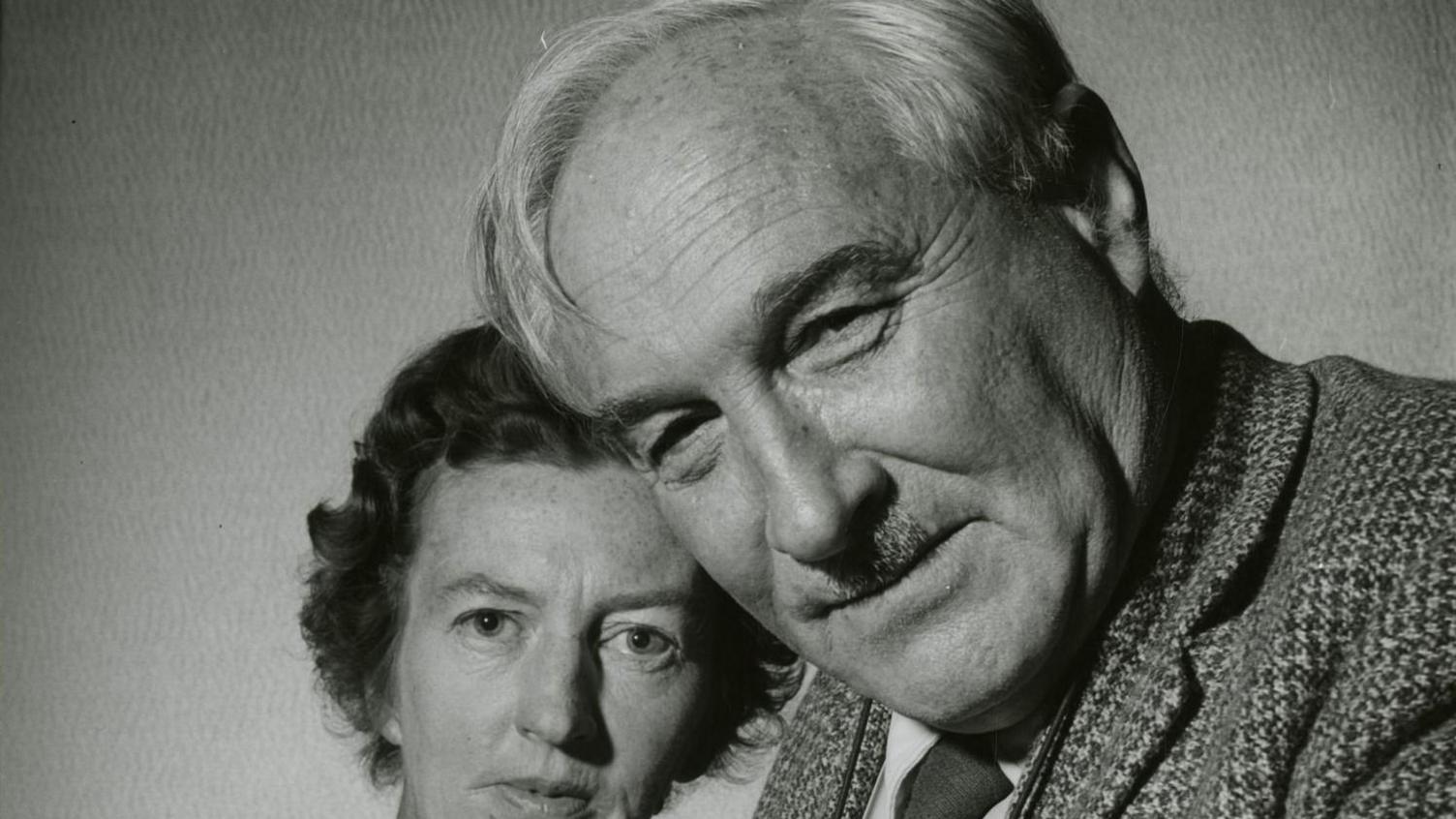 A black and white photo of Louis with his second wife Mary Leakey. He is close to the camera and has a small moustache, light coloured hair and is smiling with his mouth closed. His wife has short dark and curly hair. She further behind Louis and also looking at the camera.