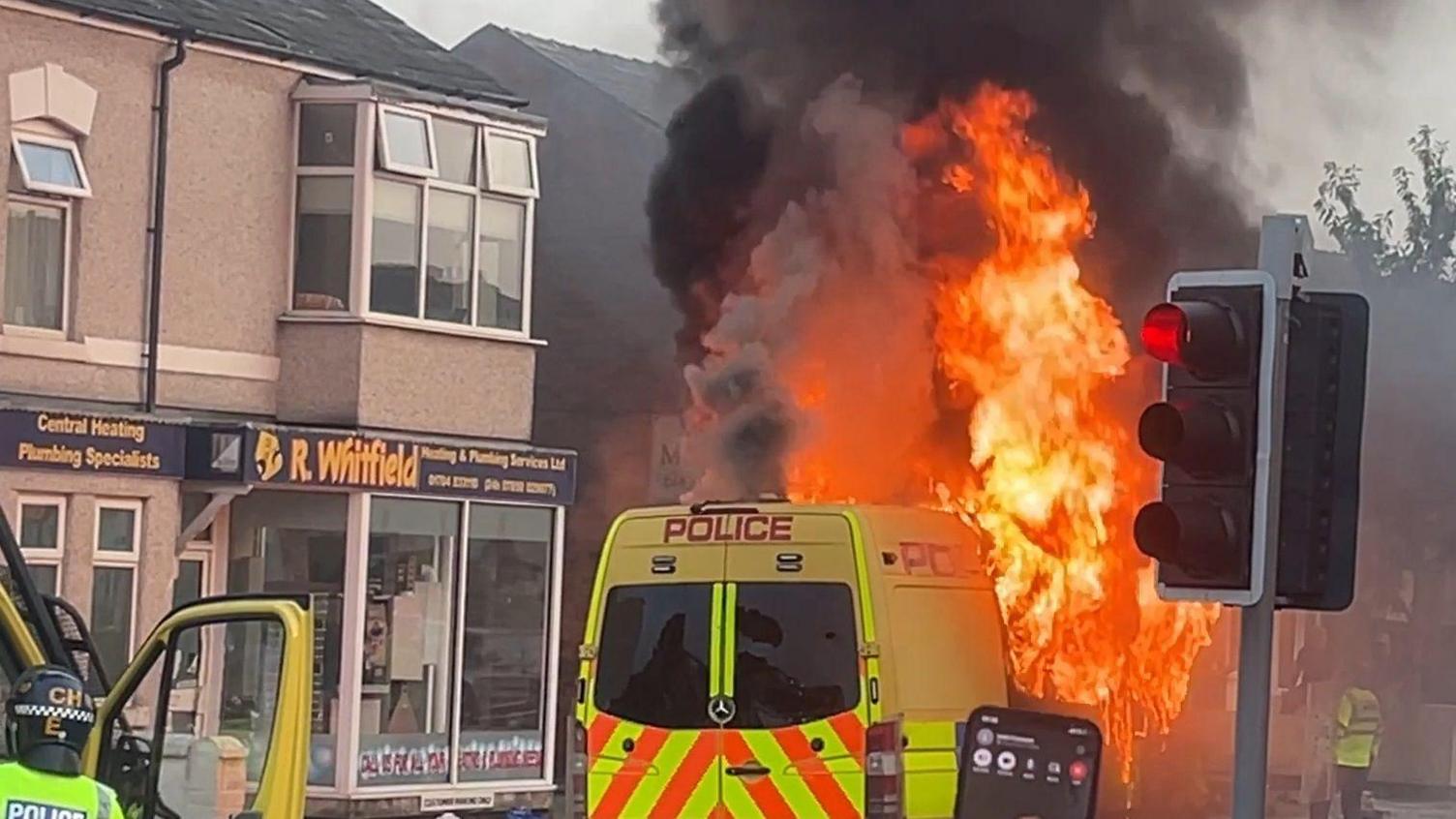 A police van on fire next to a traffic light on a road. It is parked next to a building with a sign reading R.Whitfield Central heating and plumbing specialists. It is yellow writing on black background. Another police van is parked behind it - with an officer in riot gear and door open.  