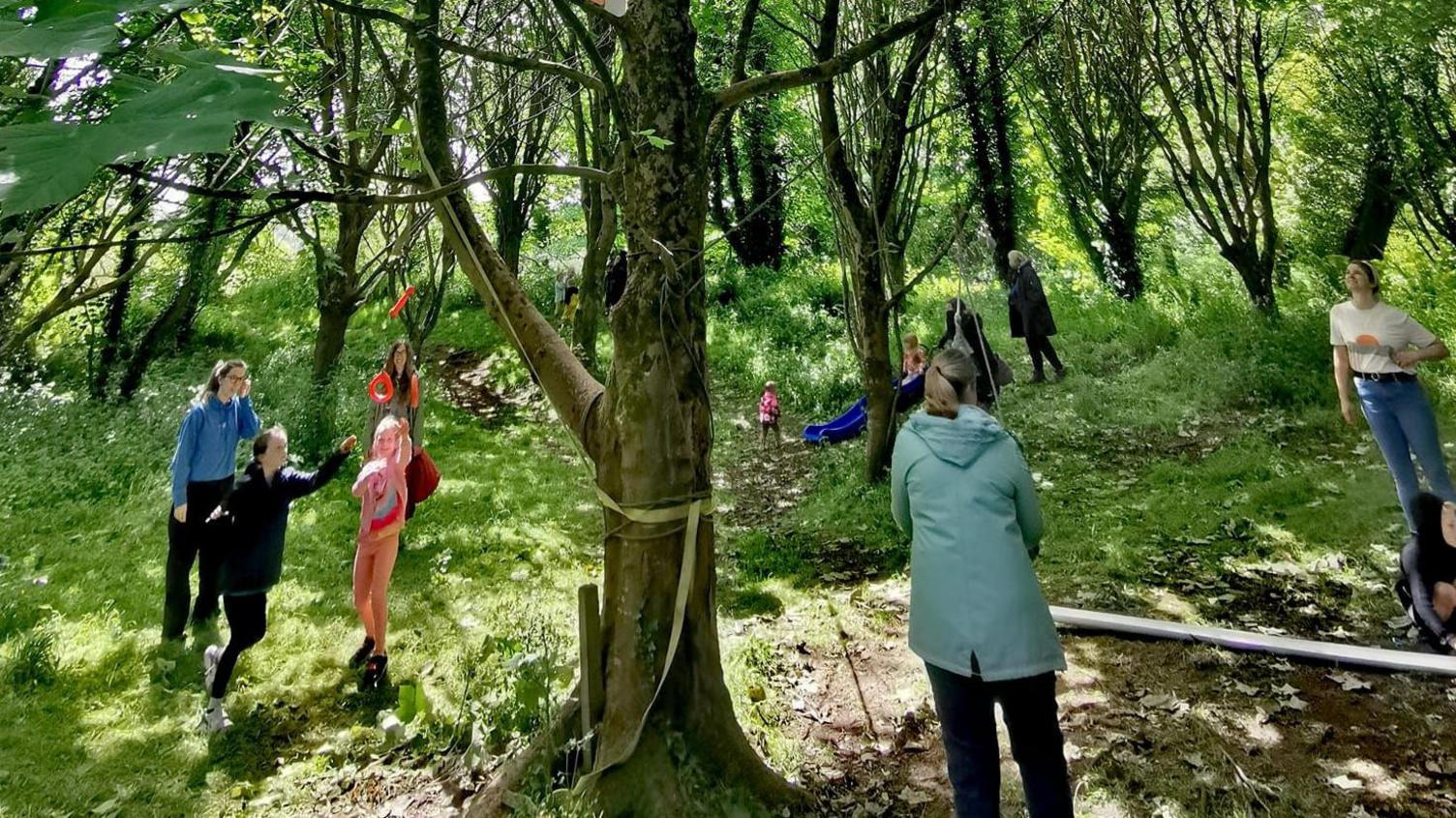 Children playing in a forest alongside adults, with the group surrounding a tree in the centre