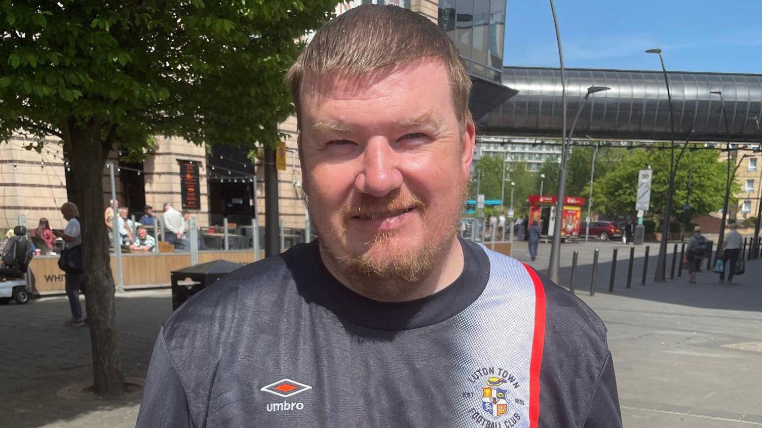 Kevin stands in the square in front of trees and a cafe. He wears a grey Luton town t-shirt with a red stripe and Umbro logo. He has short blonde hair, and a short beard and moustache.