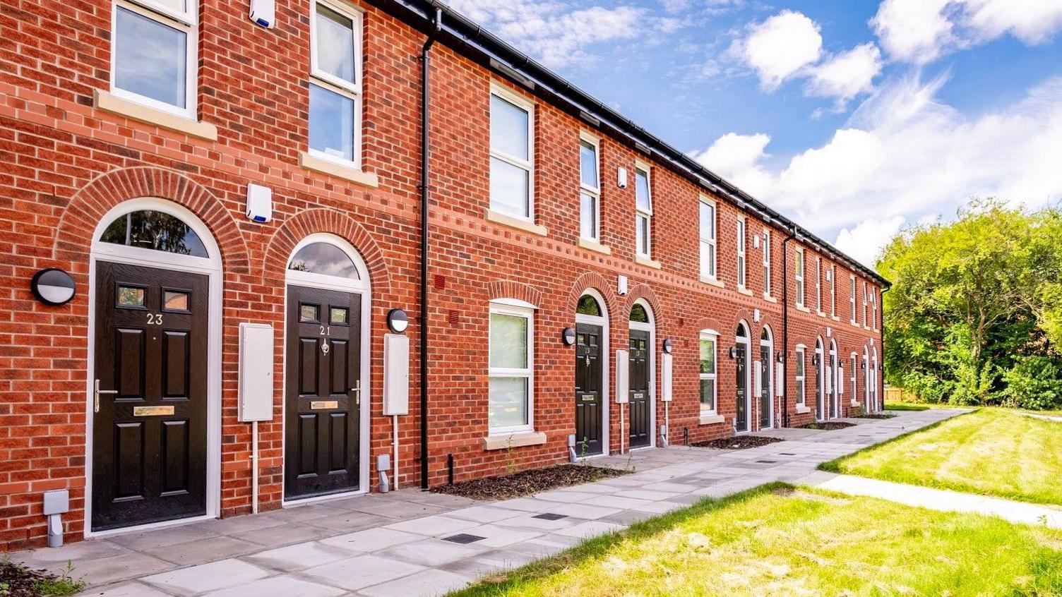 A wide shot of the front of the housing estate. It is built using red bricks and the flats all have black front doors. There are small gardens at the front.