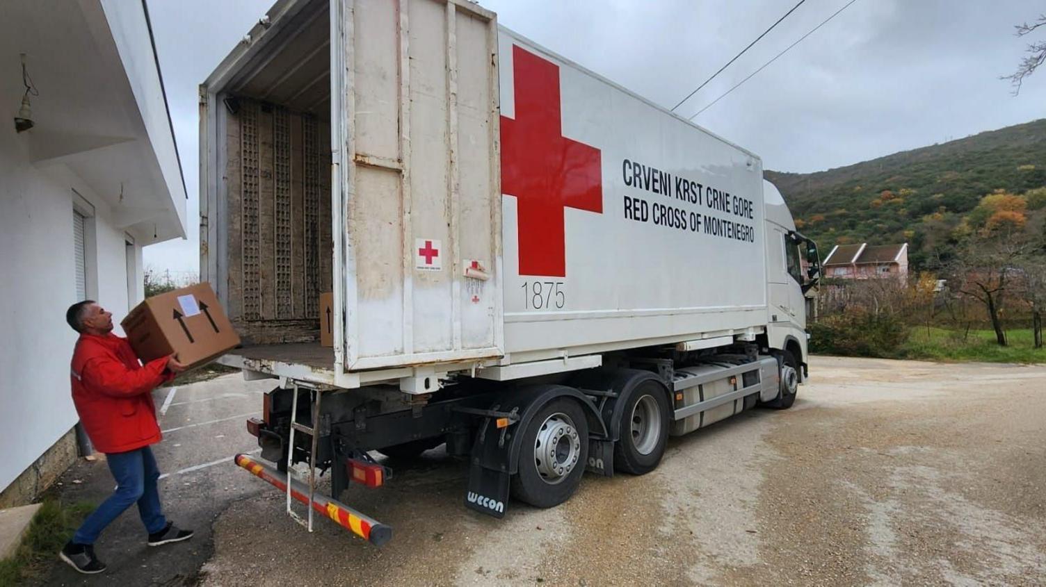 A man loads a large box onto the back of an open lorry in eastern Europe. The side of the van has a large red cross logo and reads: "RED CROSS OF MONTENEGRO" in English and Montenegrin