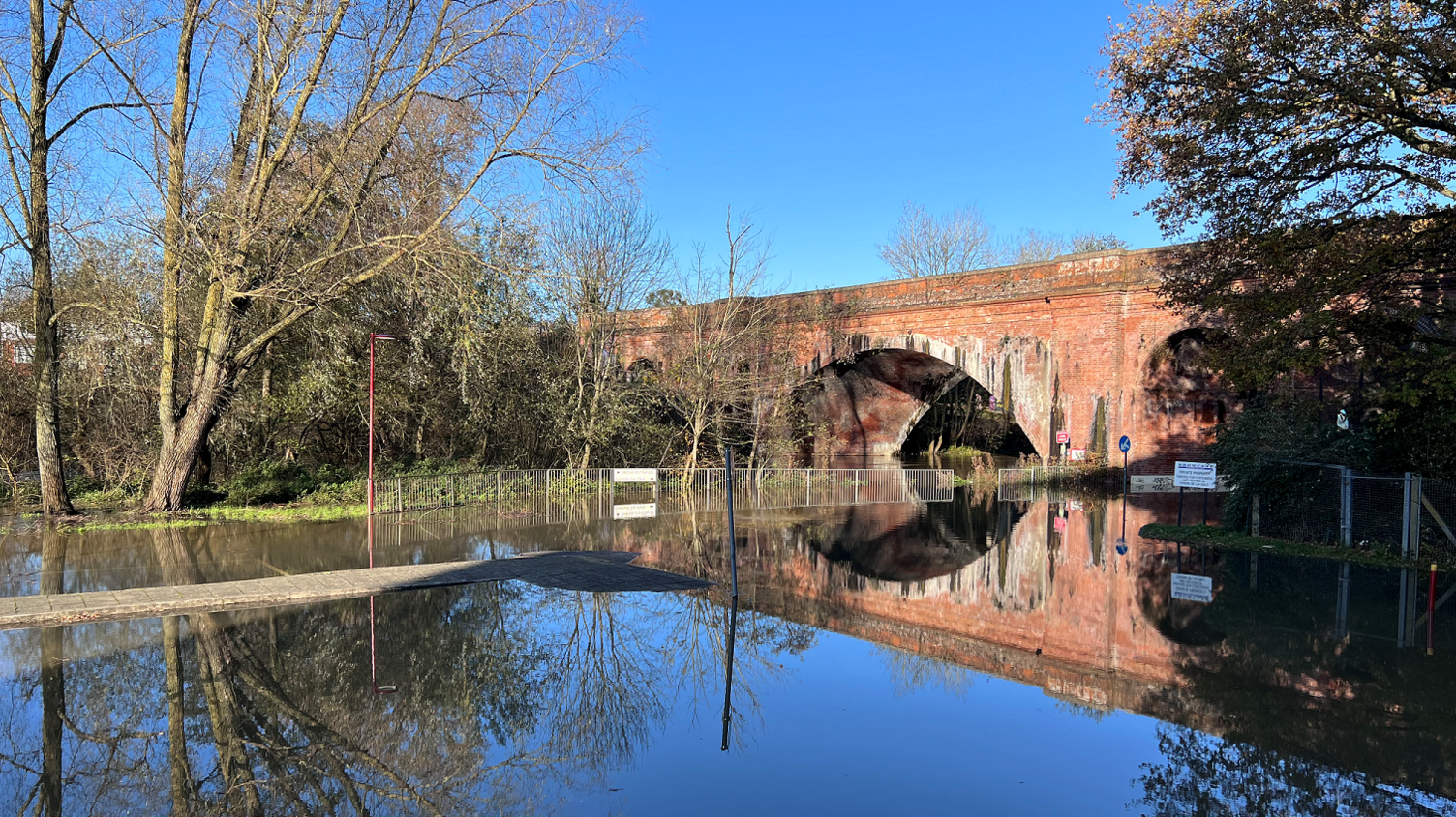 A car park covered with water, with a bridge at the back of the picture. Trees are in the distance, close to the banks of the river. 