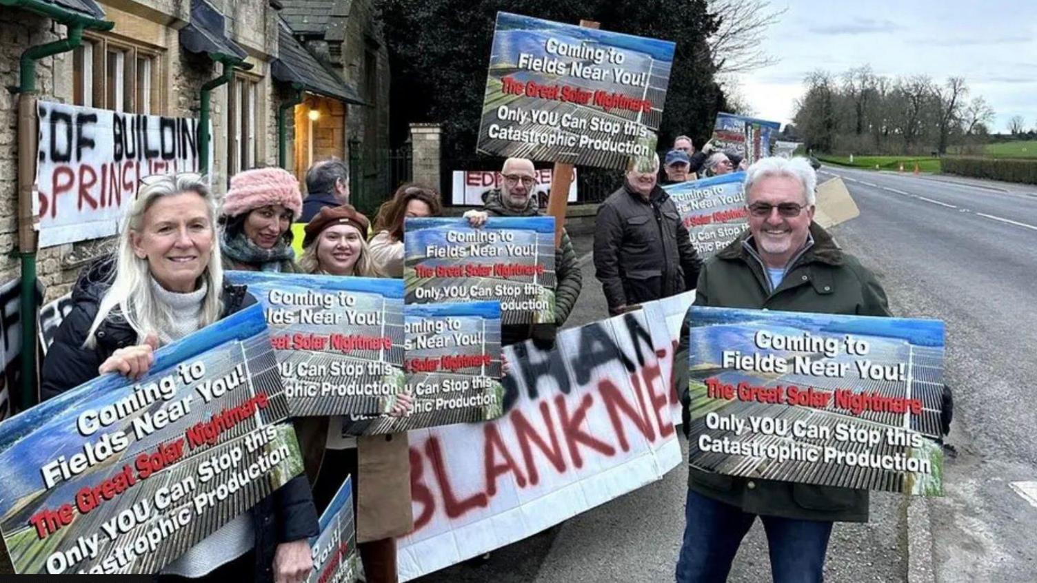 About a dozen anti solar farm campaigners hold placards. One placard reads: "Coming to fields near you. The Great Solar Nightmare. Only YOU can stop this catastrophic production". 