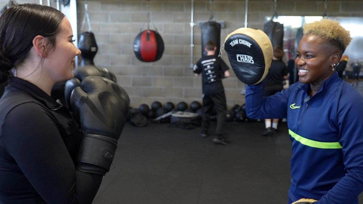 A teenage girl with black hair in a ponytail trains with Nicola, holding up her two hands inside black boxing gloves. She prepares to punch a padded cushion that Nicola is holding. Nicola wears a blue long sleeved sports top with neon strips around the middle, and has blonde short curly hair. She wears silver earrings. In the background you see other people practising with punchbags.