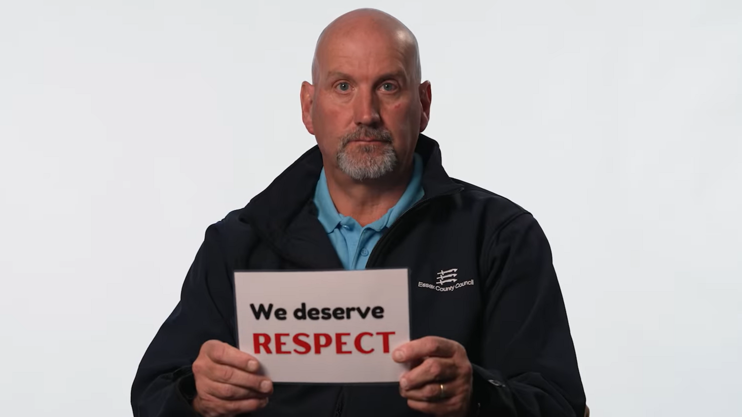 A recycling centre worker sitting in front of a white background. He has a shaved head and a goatee. He is wearing a black jumper over a blue polo top. He is holding a sign that reads "we deserve respect".