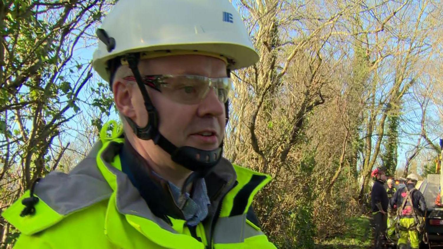 A man wearing a hard hat and a yellow high vis jacket is standing on a country road being interviewed. There are men working on repairs in the background.