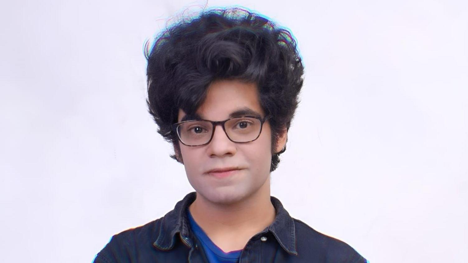 A man with a curly mop of hair and glasses stands in front of a white background. He wears a denim jacket over a dark blue t-shirt.