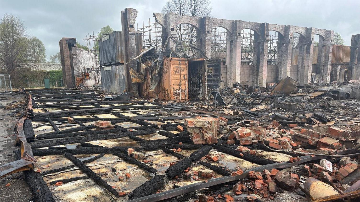 Burned debris and bricks with large brick archways in the background. A burned-out shipping container can be seen. Grey skies.