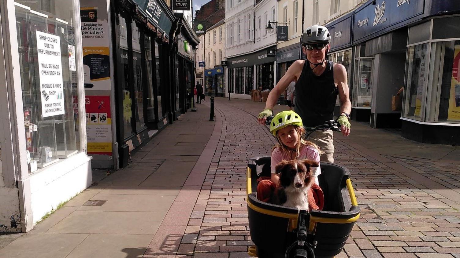 A man cycling along a cobbled street, with a girl and dog in a wagon attached to the front of the bike.