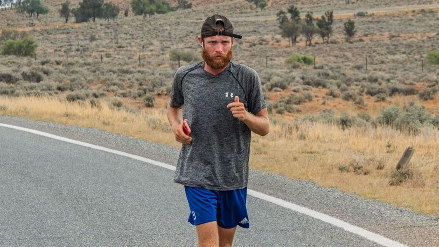 Jack Pitcher runs along a road. He has a ginger beard and wears a baseball cap back-to-front, a grey T-shirt and blue football shorts.