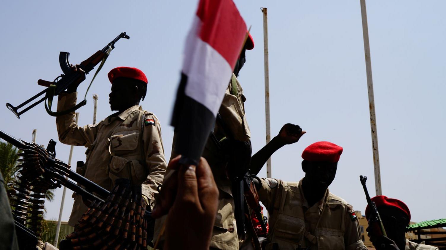 Fighters from the Rapid Support Forces raise their fists and guns in the air. They are wearing red berets and military clothing.