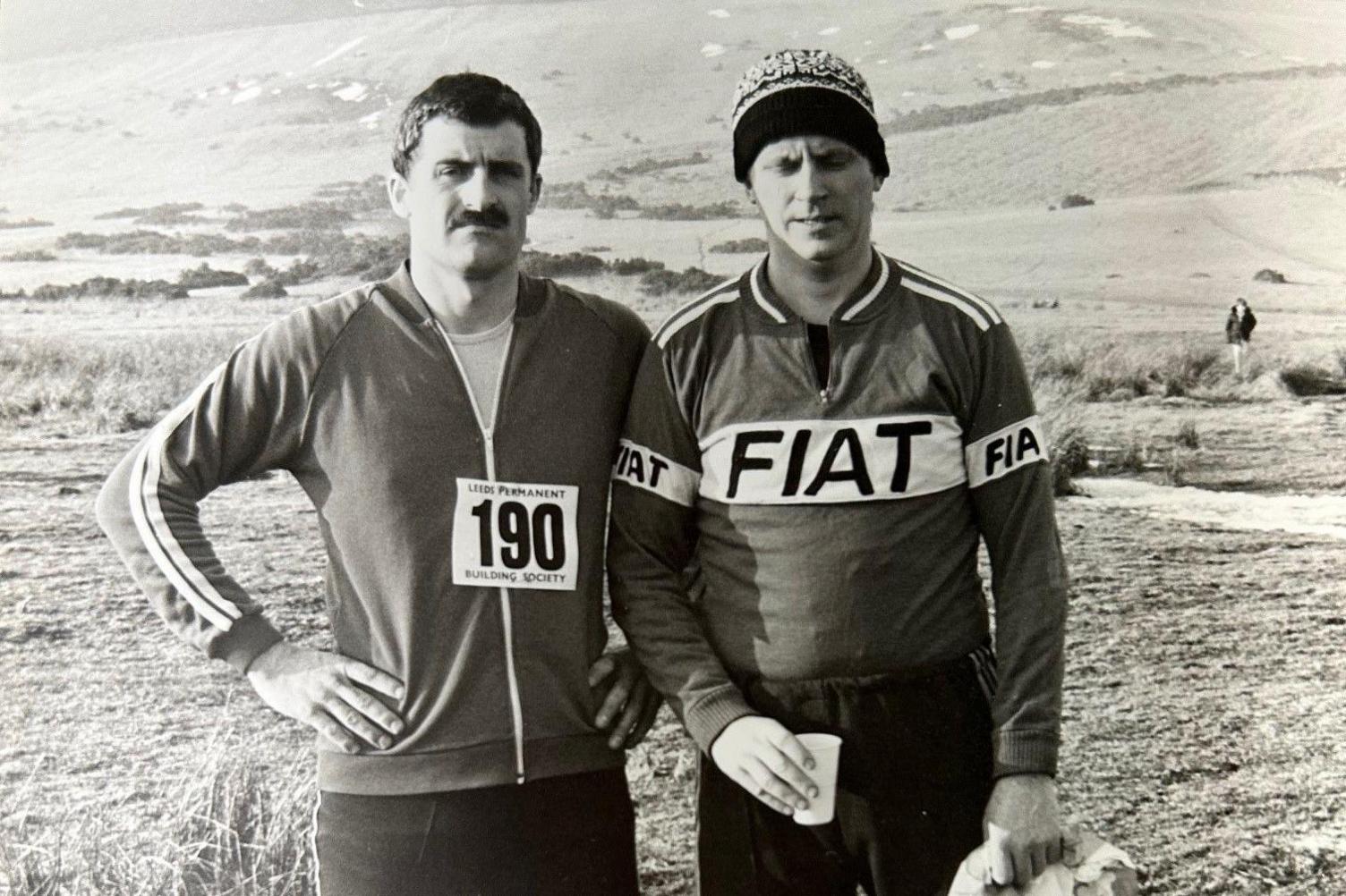 A black and white picture of Mr Allan and Mr Anderson in running clothes surrounded by hills. Mr Anderson is wearing a woolly hat. Mr Allan has a moustache.