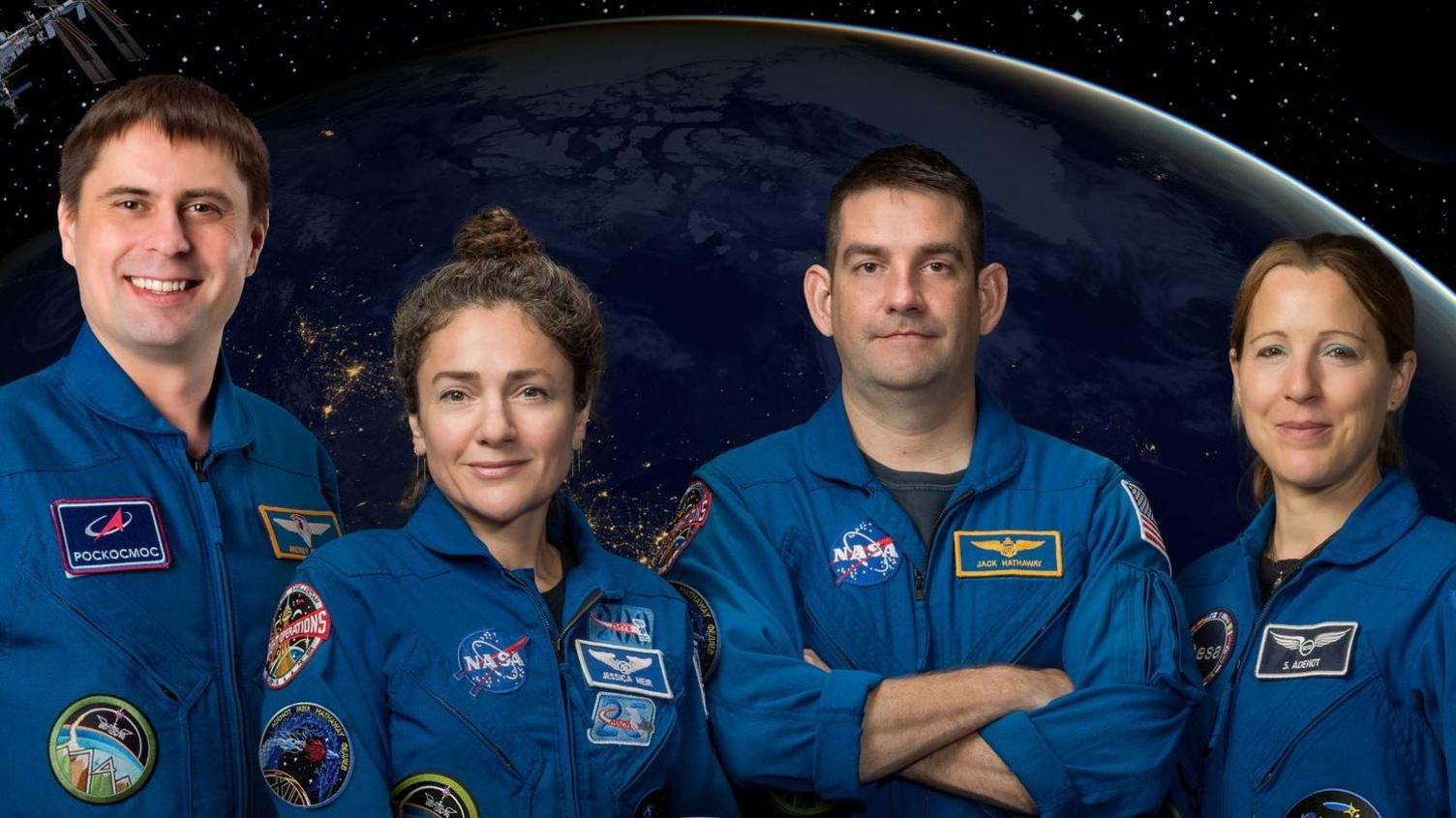 Two men and two women wearing blue overalls covered in badges, including the NASA logo. They're stood in front of a graphic which shows the view of the Earth from space
