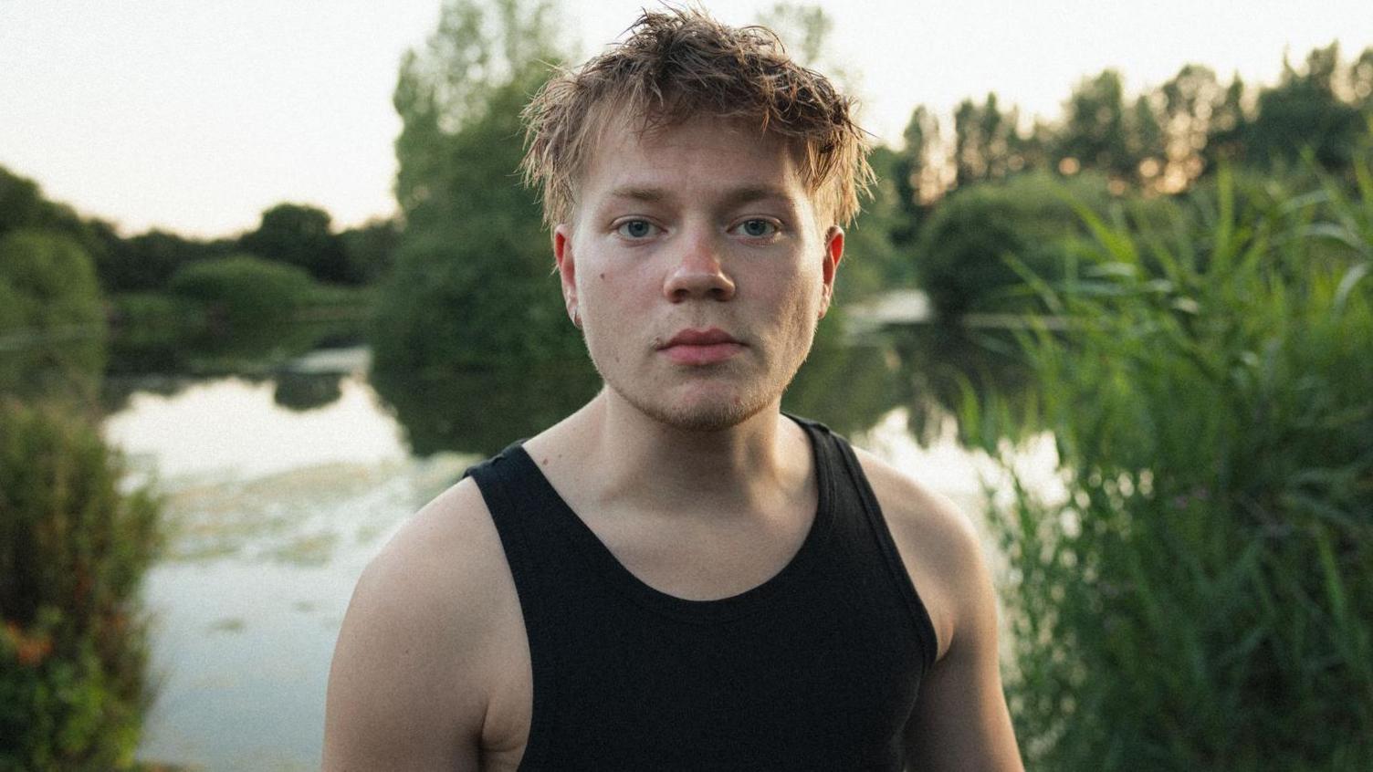 A man in a black sleeveless top stands, with a serious expression. He is stood in front of a lake, surrounded by green trees and foliage. The water reflects the natural scenery.