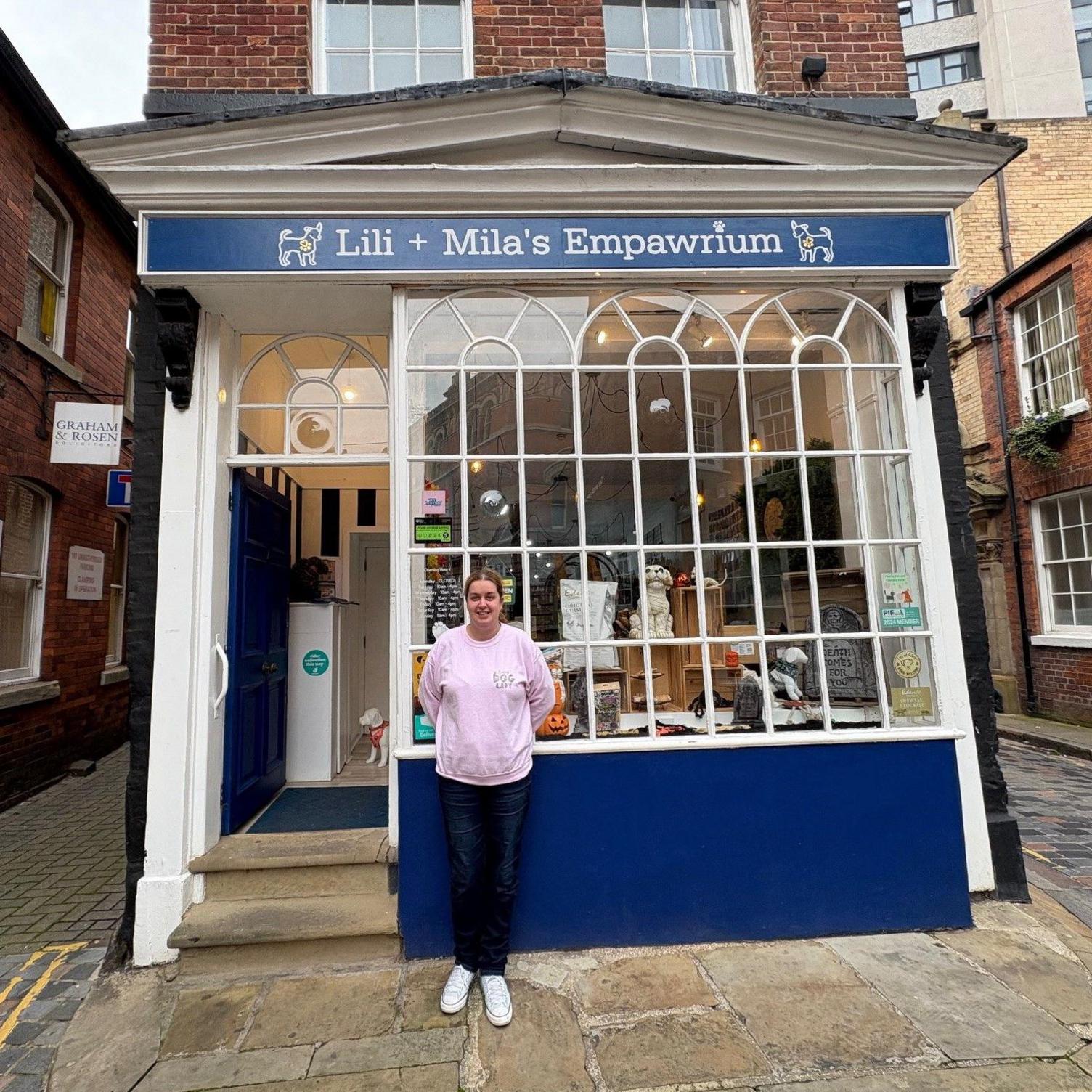 A woman in her 20s is standing in front of her pet shop which is painted blue. It has a large glass frontage containing panes.