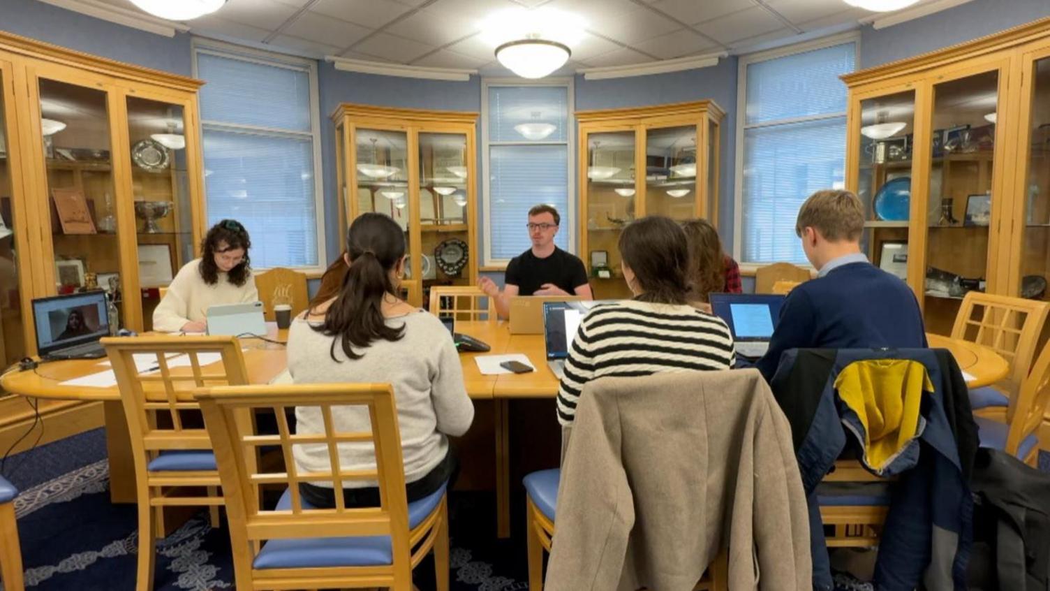 Seven young people in the Youth Select Committee sit around a long oval table in a curved office, with one member visible on a laptop joining remotely. The office has large glass cabinets lining the walls, broken up by three tall windows. The furniture is all pine wood, and the walls and seats are in a pale blue colour.