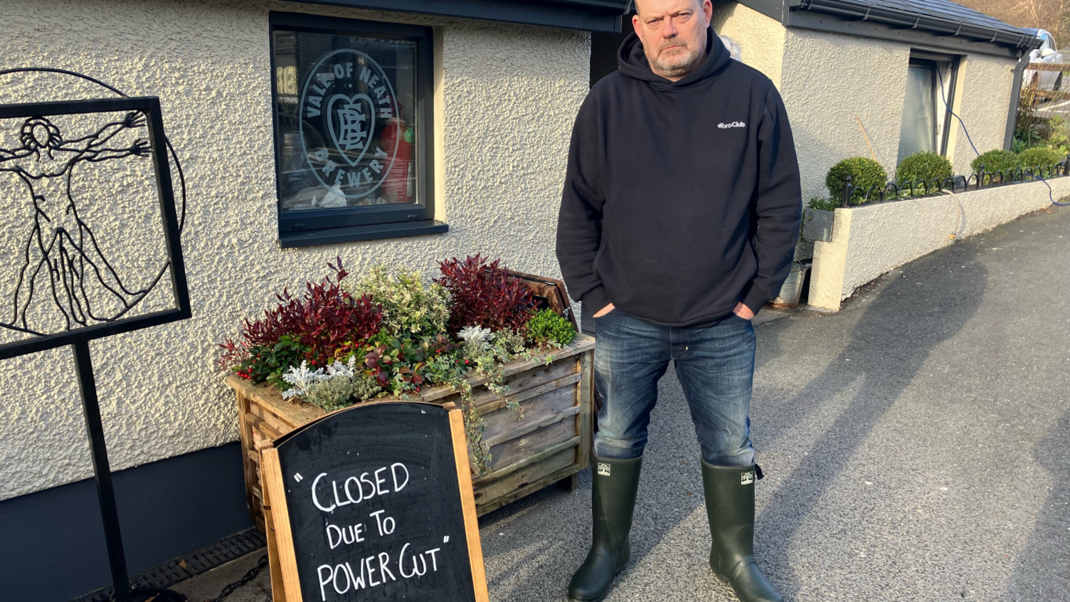 Nils Kirk, wearing wellington boots, jeans and a dark sweatshirt, standing outside his pub alongside a sign reading 'Closed due to power cut'