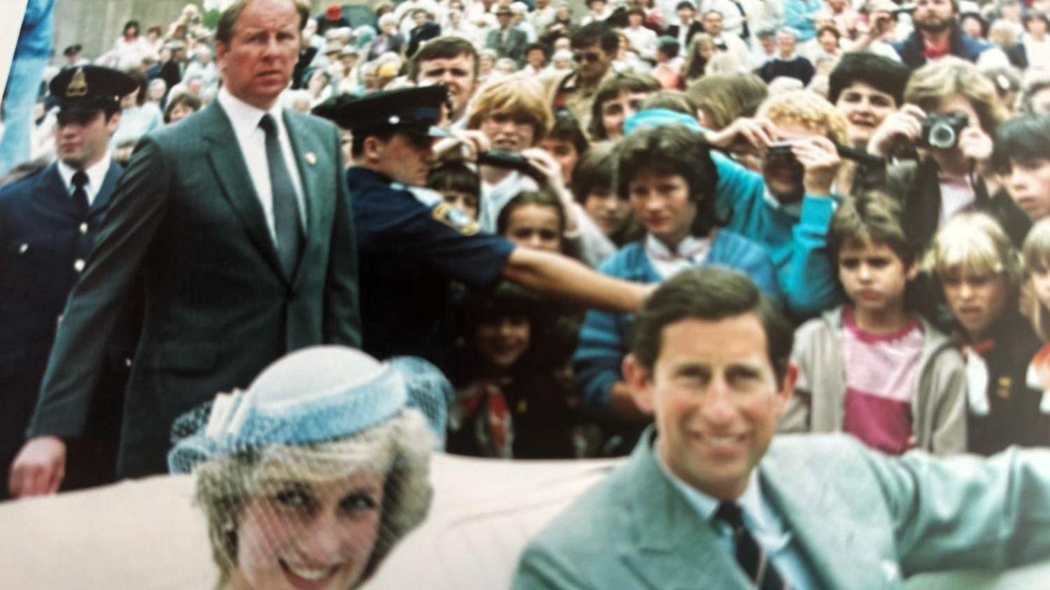 Princess Diana and then-Prince Charles sit in a white car in the foreground of the photo. Prince Charles is wearing a grey suit and Princess DIana has on a blue fascinator. In the background, a man in a dark suit can be seen - this is Allan Peters.