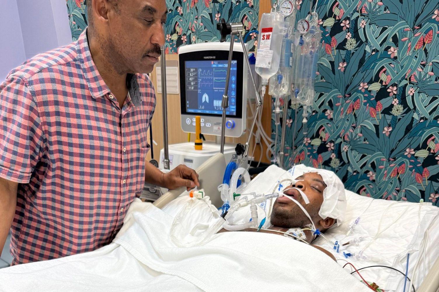 Okemena Sule in hospital surrounded tubes, medical drips and monitors as a family member watches on