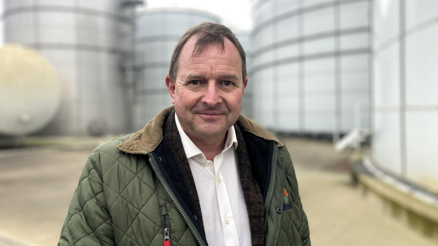 Lord John Fuller wearing a white shirt and green jacket smiling while stood in front of fertiliser silos