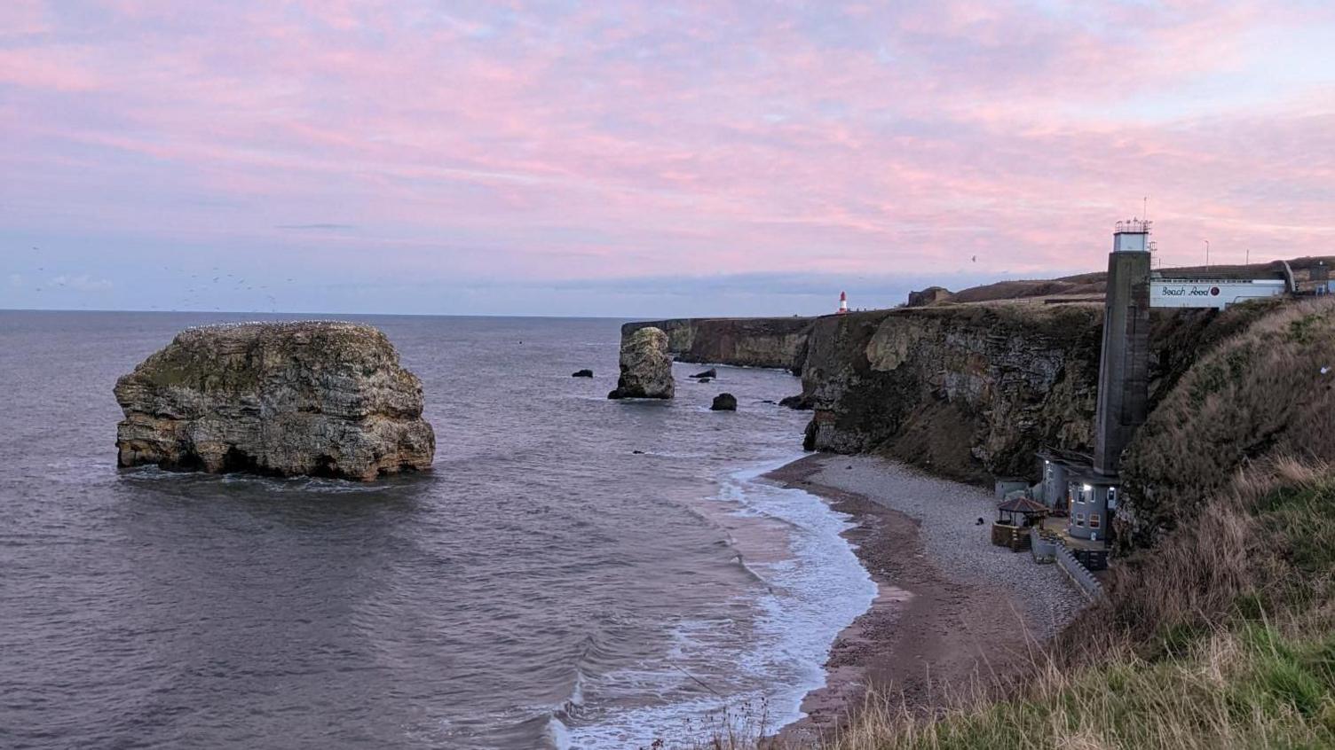 Purple clouds over Marsden Rock, a large rock surrounded by the sea. It is a short way away from a beach, where there is a restaurant at the bottom with a large concrete lift shaft leading to the top