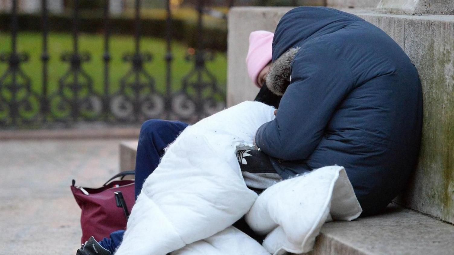 A rough sleeper is pictured sitting on a stone wall clutching a duvet, wearing a navy coat and pink beanie. Their head is nestled into the duvet.