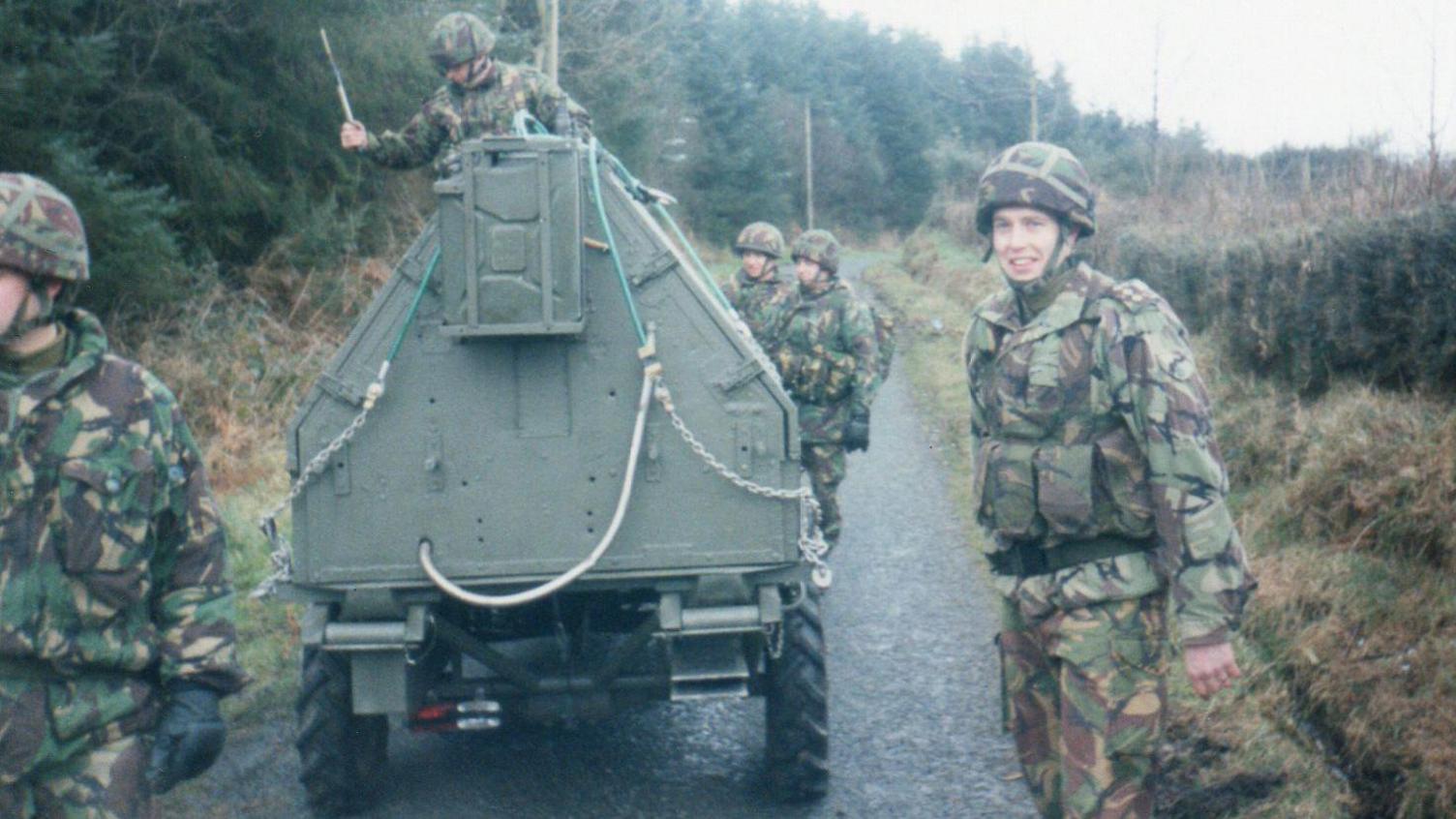 Gareth is one of five soldiers searching for IRA roadside devices. Dressed in army uniform, they are walking down a narrow country road with pine trees to the left and a hedgerow to the right