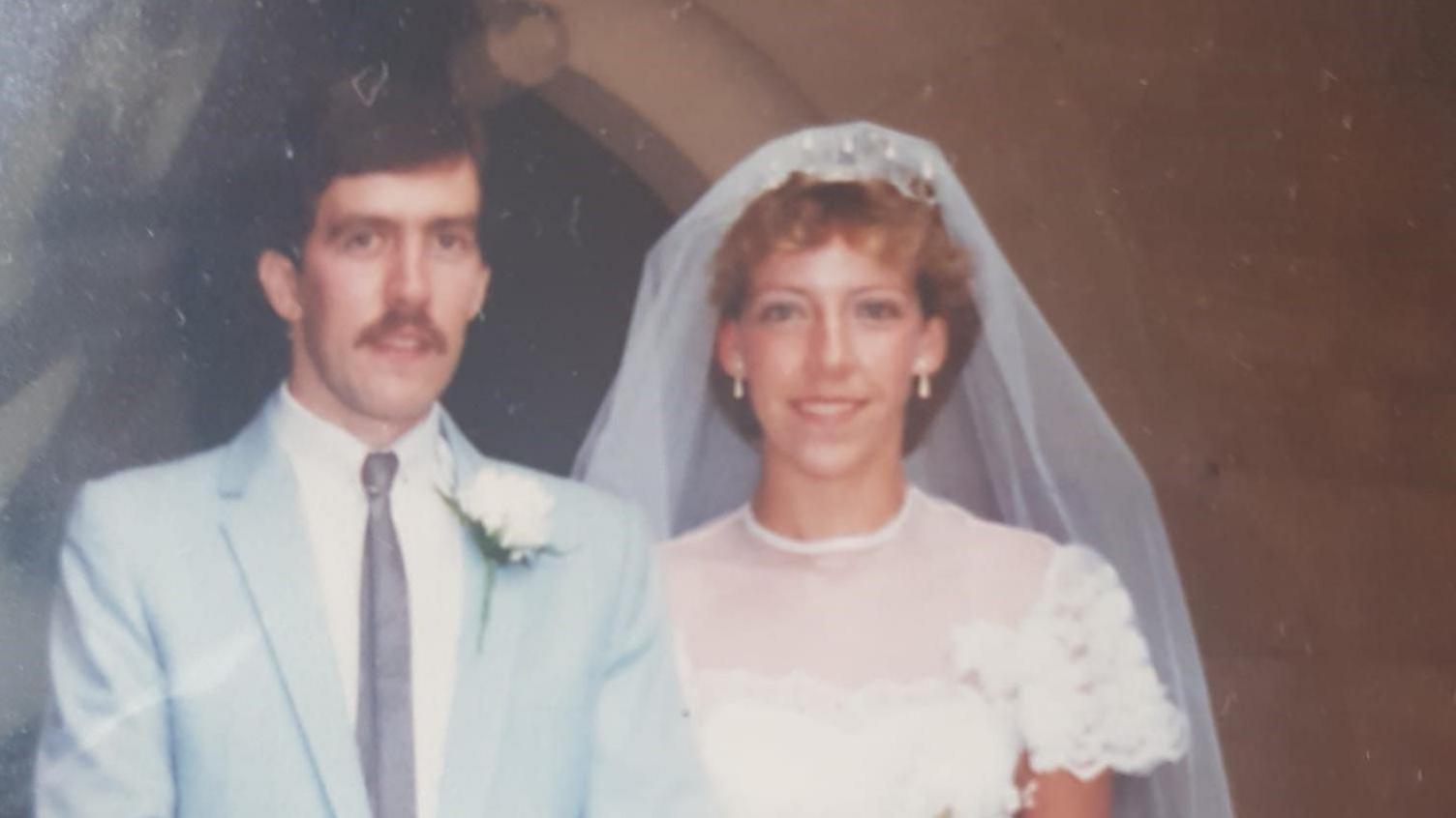 A married couple in the 1980s. The groom is wearing a pale blue suit, white rosette and moustache. The bride has blonde hair and is wearing a white dress with a veil.