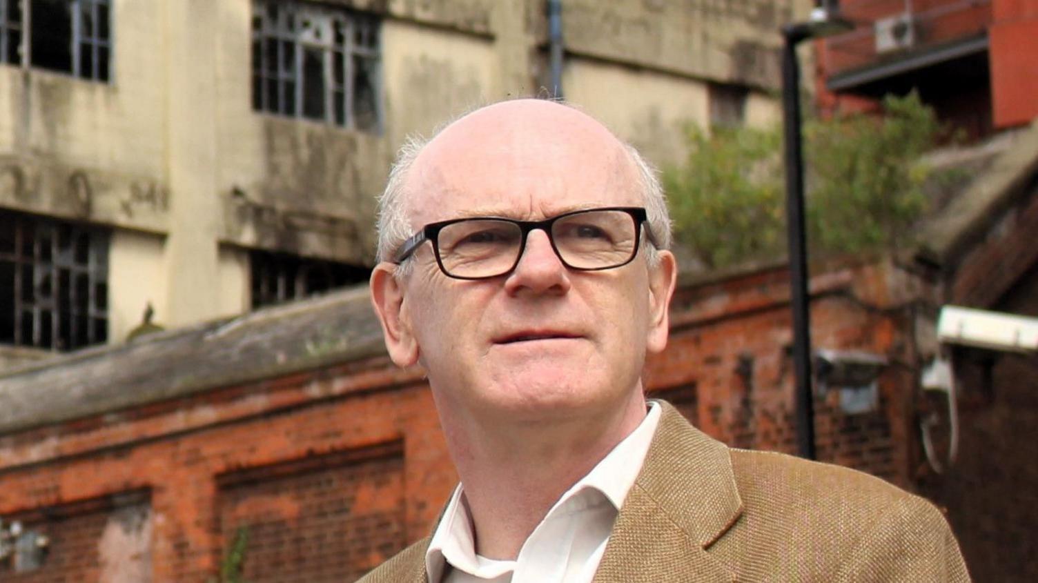 Neil Macdonald with black glasses looks away from the camera. He wears a light brown suit jacket and white shirt underneath.