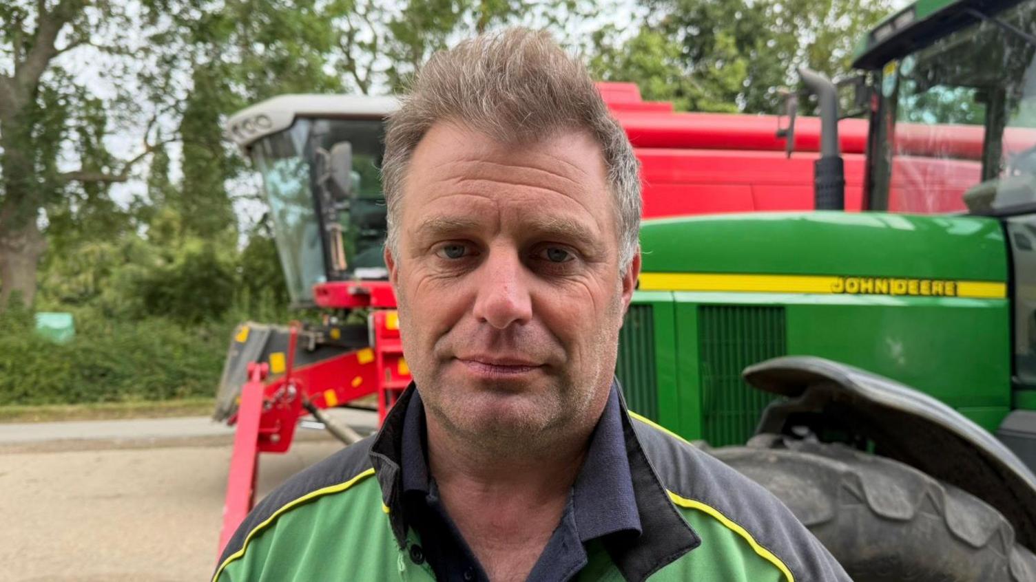 Henry Moreton looking at the camera. He is pulling a straight face, and is wearing green and blue farming overalls. Behind him is a green John Deere tractor, and a red combine harvester.