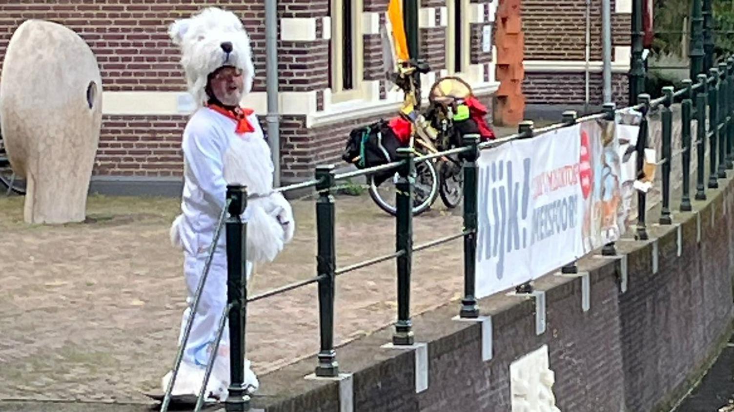 A man in a polar bear costume standing behind railings next to ta canal. He is squirting liquid into the canal - using a mechanical device to simulate urination.