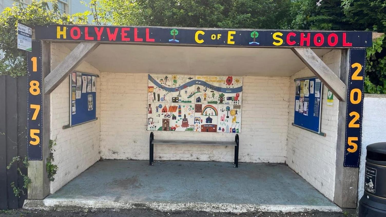 A brick built white washed bus shelter 4m (12ft) long and 3m (9ft) deep decorated with paintings and writing about the Holywell C of E Primary school.