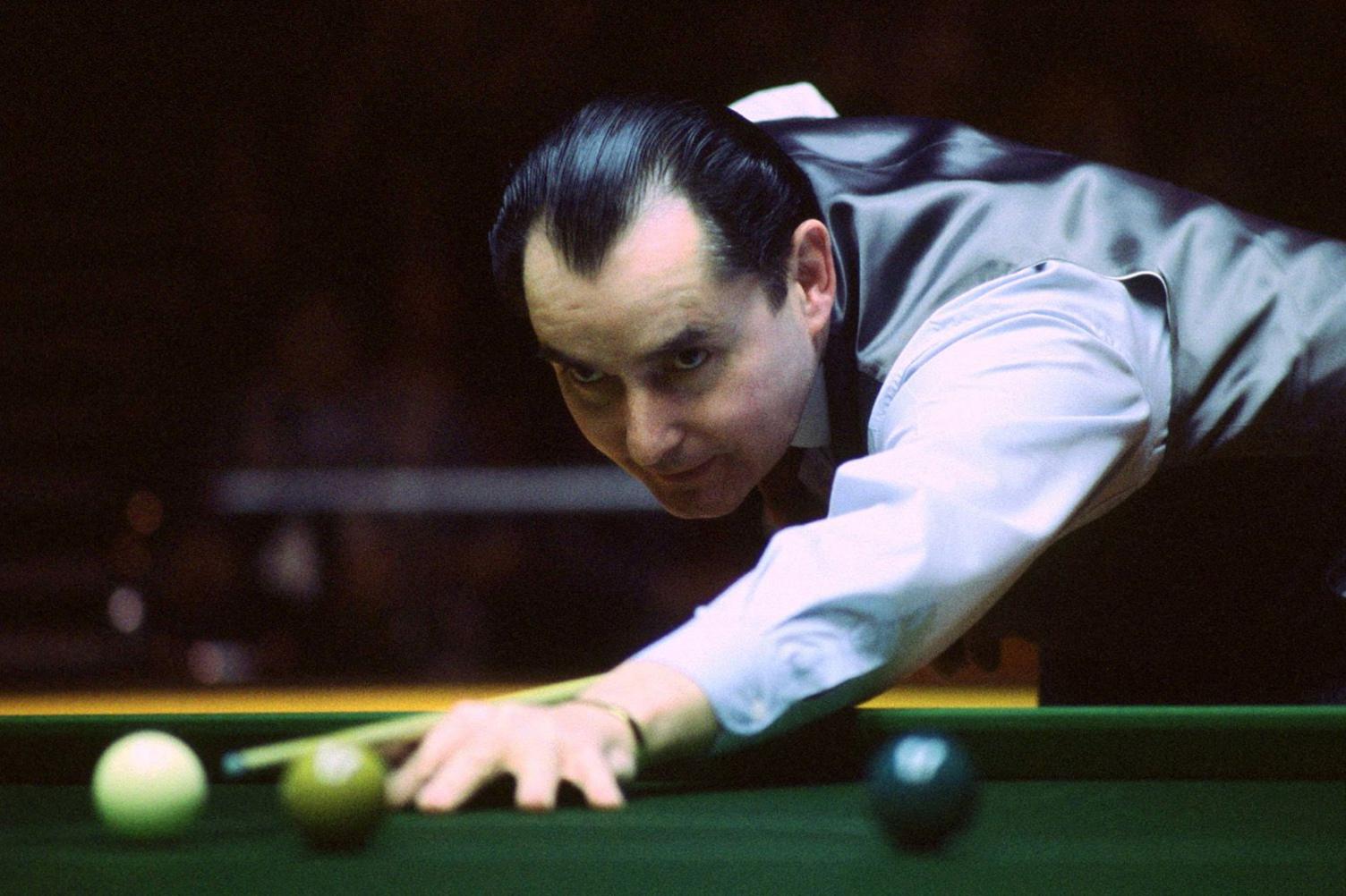 Ray Reardon, pictured in his playing days. He is leaning over the snooker table, getting ready to hit the white ball. He is wearing a white shirt and waistcoat favoured by nearly all snooker players. His hair is very dark and slicked back into a "widow's peak"