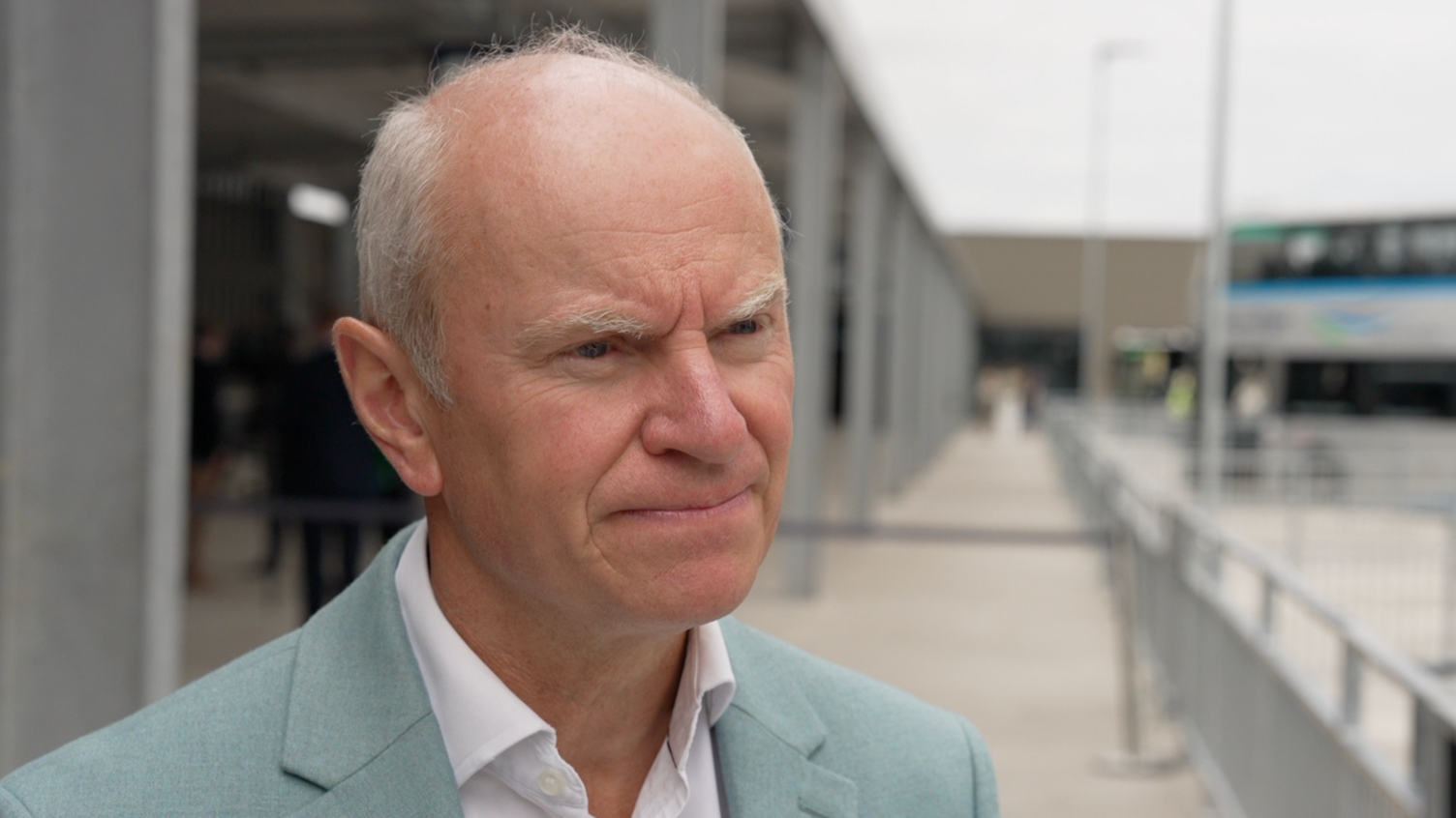 Dave Lees, who wears a light green suit and white shirt, looks on with a serious expression while being interviewed.